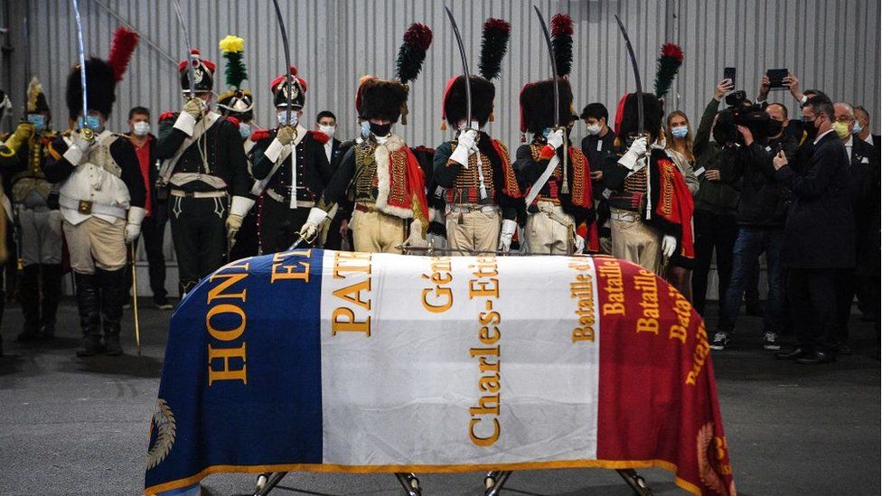 Men dressed as Napoleon-era fighters stand in respect beside a French flag-covered coffin